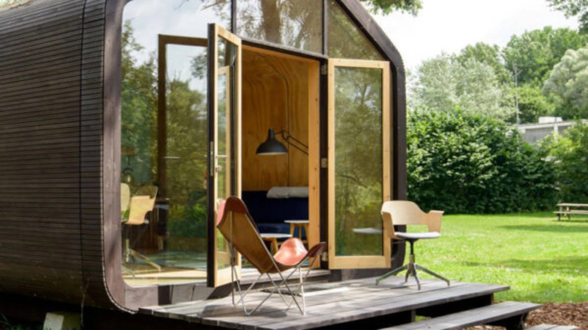 Pequeña vivienda modular de diseño minimalista con fachada oscura, grandes ventanales de vidrio y terraza de madera rodeada de jardín verde.