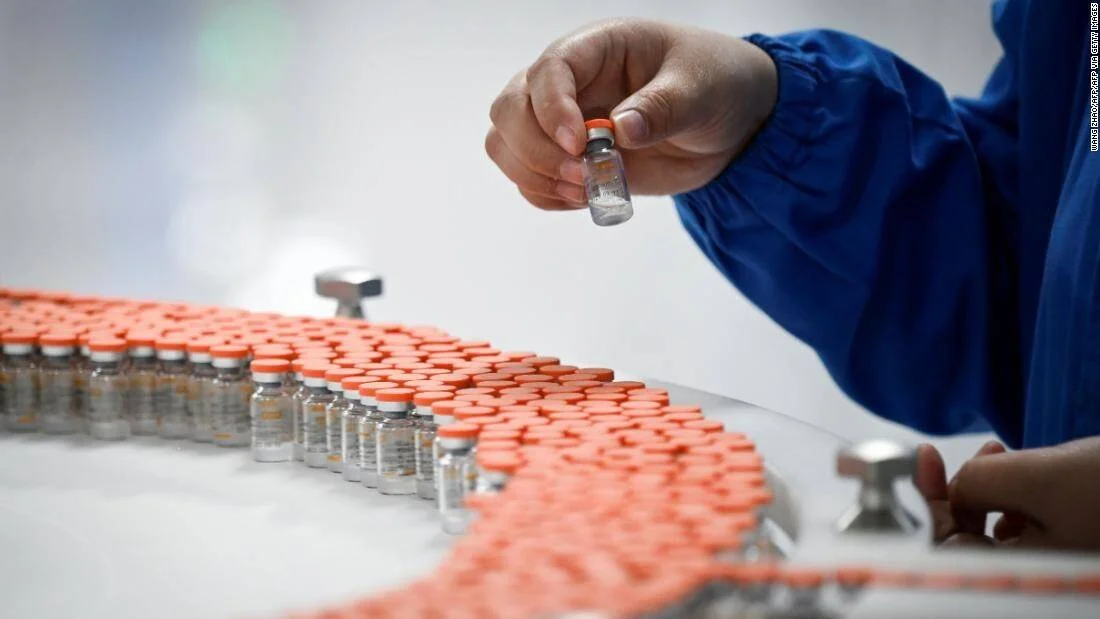 Trabajador con bata azul sostiene un vial de vacuna sobre una línea de producción con cientos de frascos de tapa naranja.