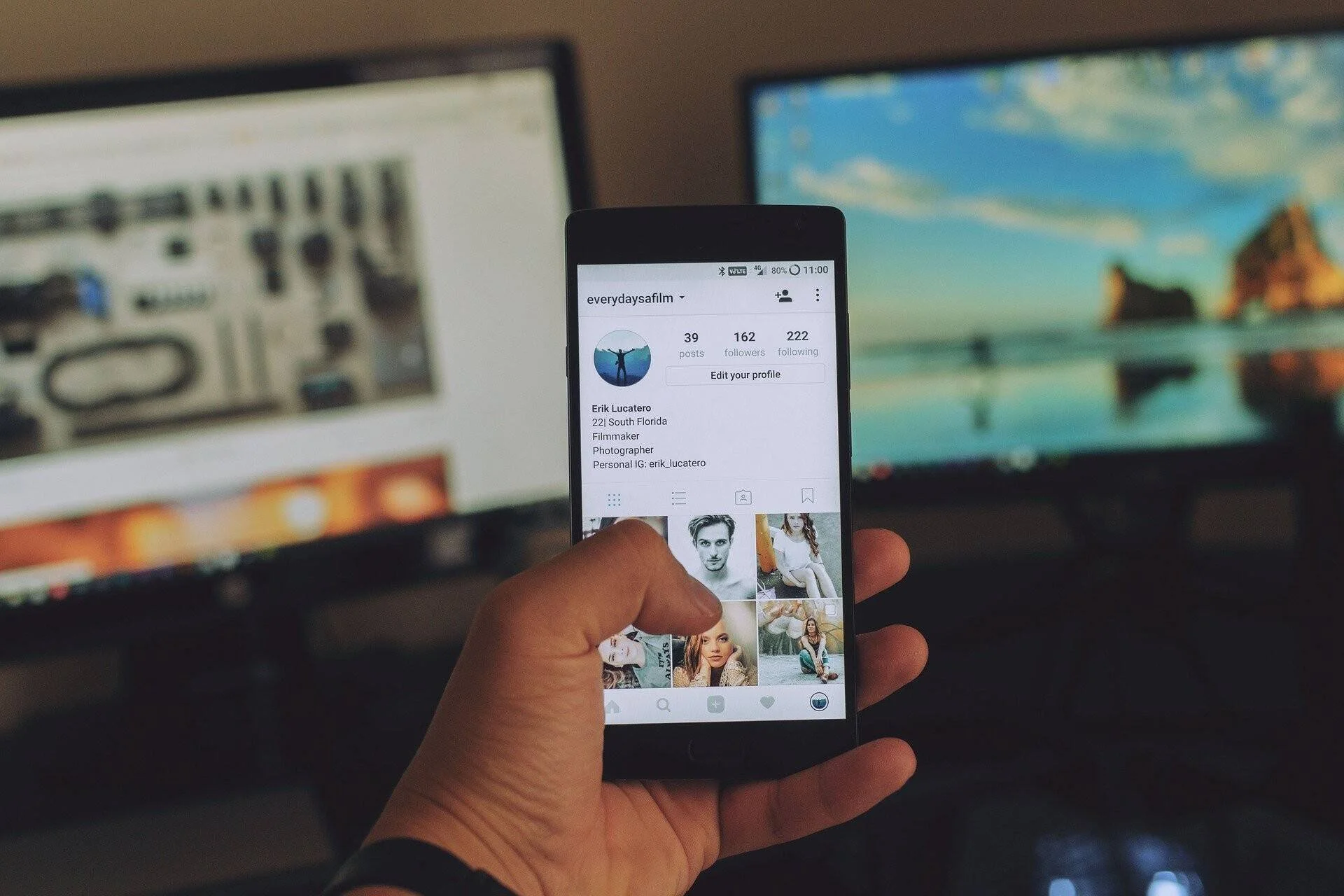 Una mano sostiene un teléfono con un perfil de Instagram abierto, con monitores de computadora al fondo.