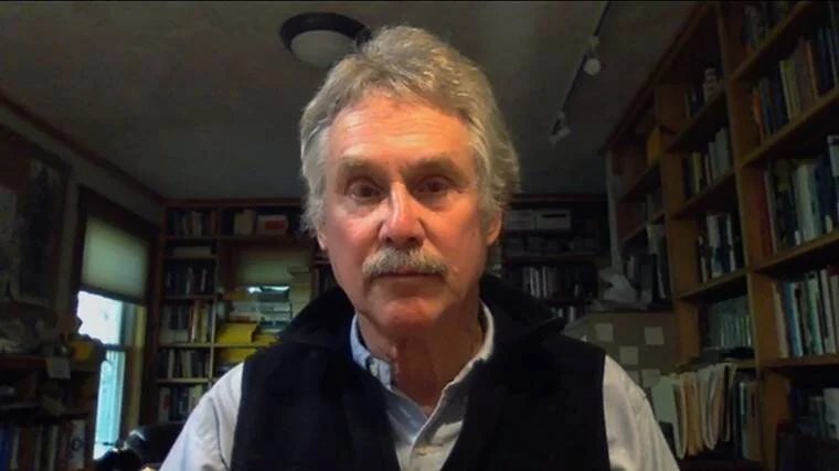 Hombre de cabello canoso y bigote gris habla frente a una cámara con estanterías llenas de libros al fondo.