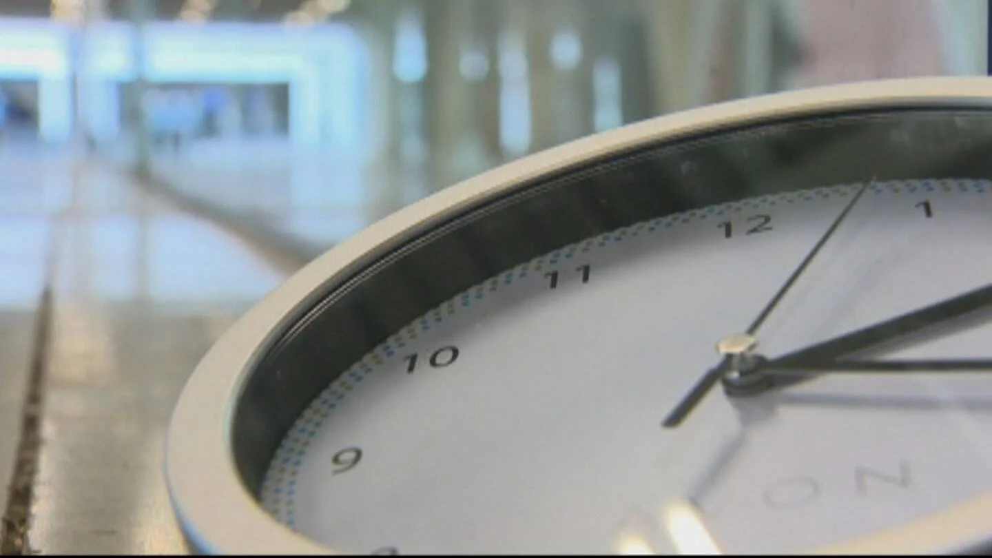 Reloj de pared analógico que marca aproximadamente las 12:58, con un edificio moderno desenfocado al fondo.
