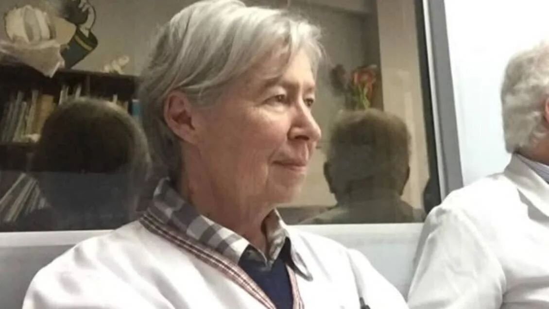 Mujer de cabello gris con guardapolvo blanco sentada en un entorno médico, mirando hacia un lado.