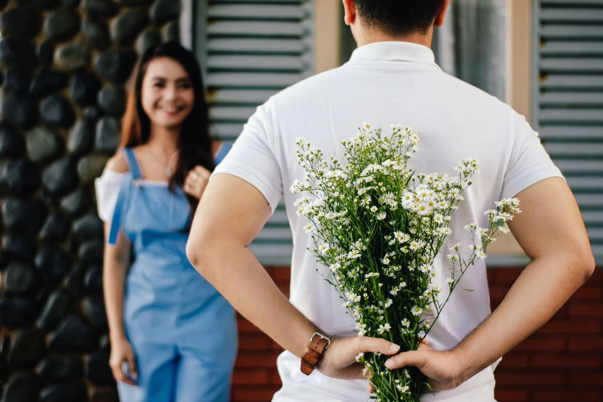 Un hombre esconde un ramo de flores blancas detrás de su espalda frente a una mujer sonriente con overol azul.