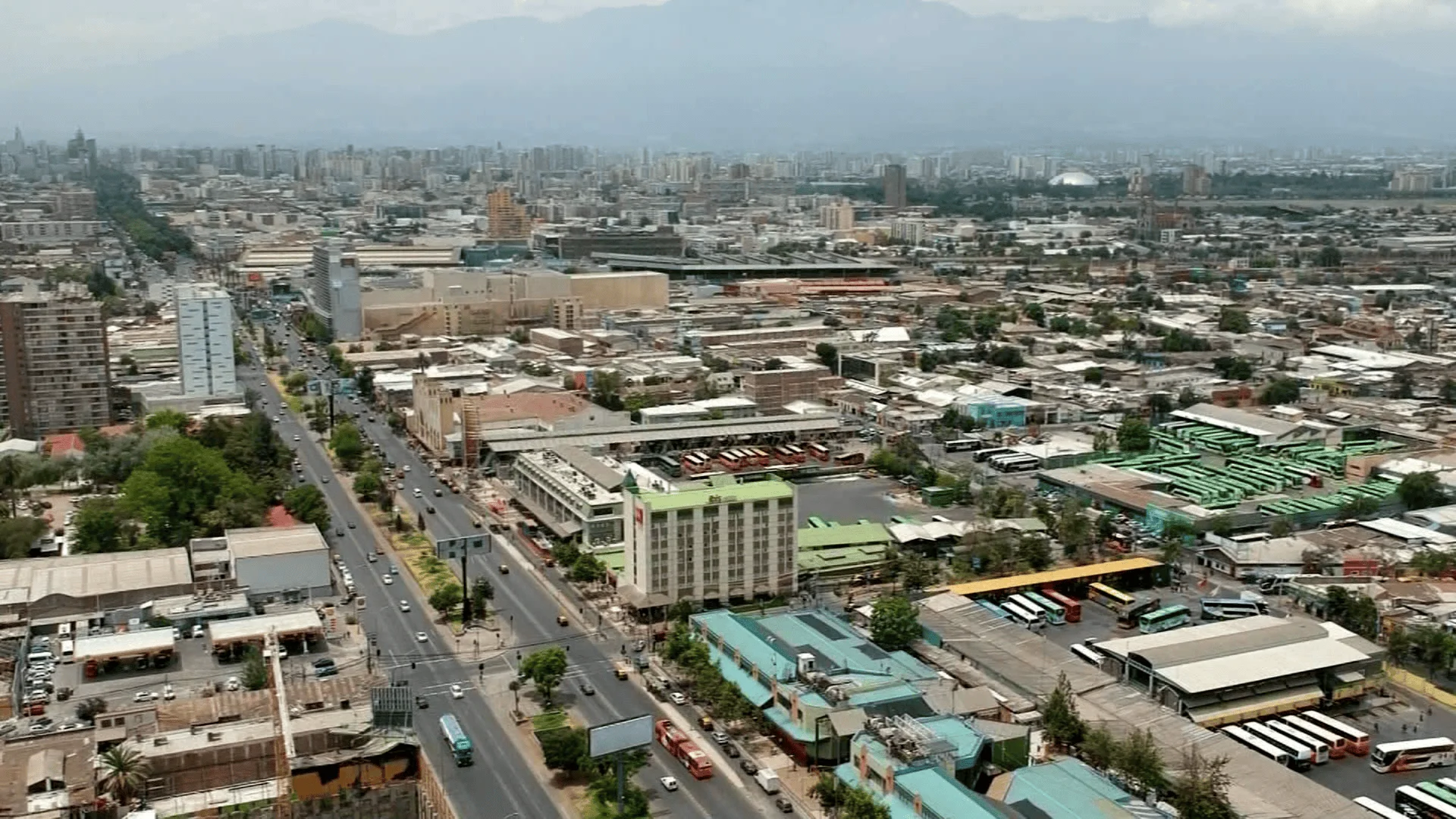 Panorámica aérea de una ciudad con una gran avenida, un terminal de buses y edificios con la cordillera al fondo.