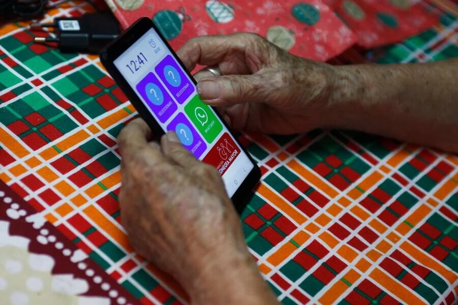 Manos de una persona mayor sostienen un celular con pantalla de botones grandes y coloridos, incluido WhatsApp.