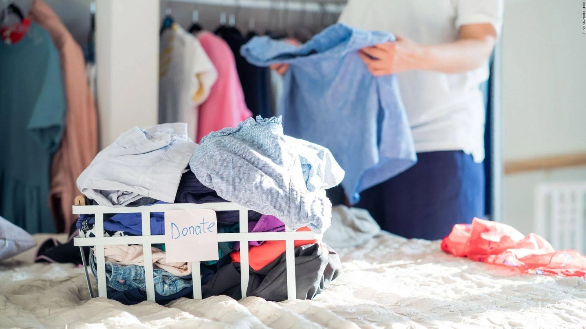 Persona seleccionando ropa frente a un armario, con una cesta etiquetada 'Donate' llena de prendas sobre la cama.