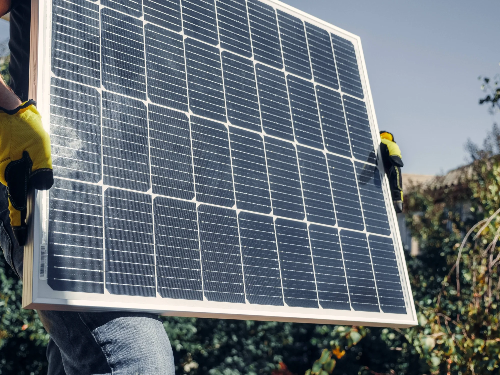 Un trabajador con guantes amarillos carga un panel solar fotovoltaico al aire libre durante su instalación.