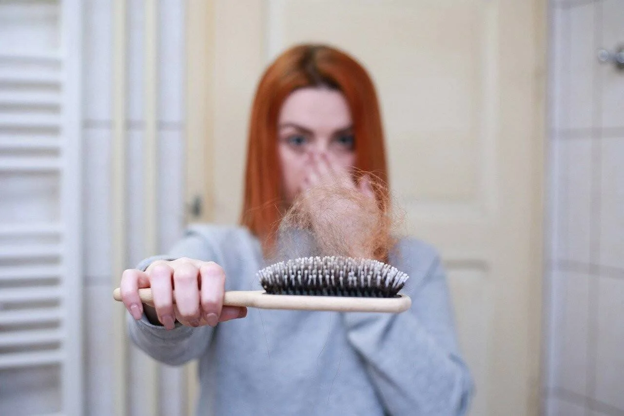Mujer pelirroja sostiene un cepillo lleno de cabello caído, con gesto de preocupación tapándose la boca.