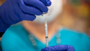 Manos con guantes azules cargan una jeringa extrayendo líquido de un vial de vacuna.