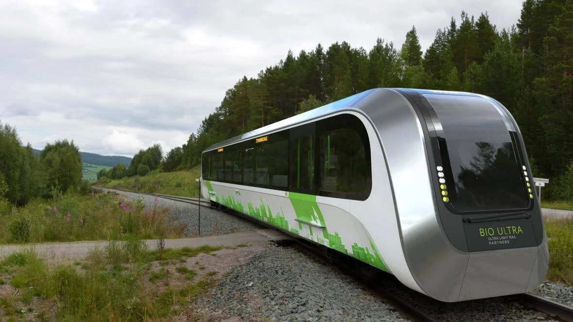 Tren ultraligero Bio Ultra Partners de diseño futurista plateado y verde circula por vía rural rodeada de bosque.