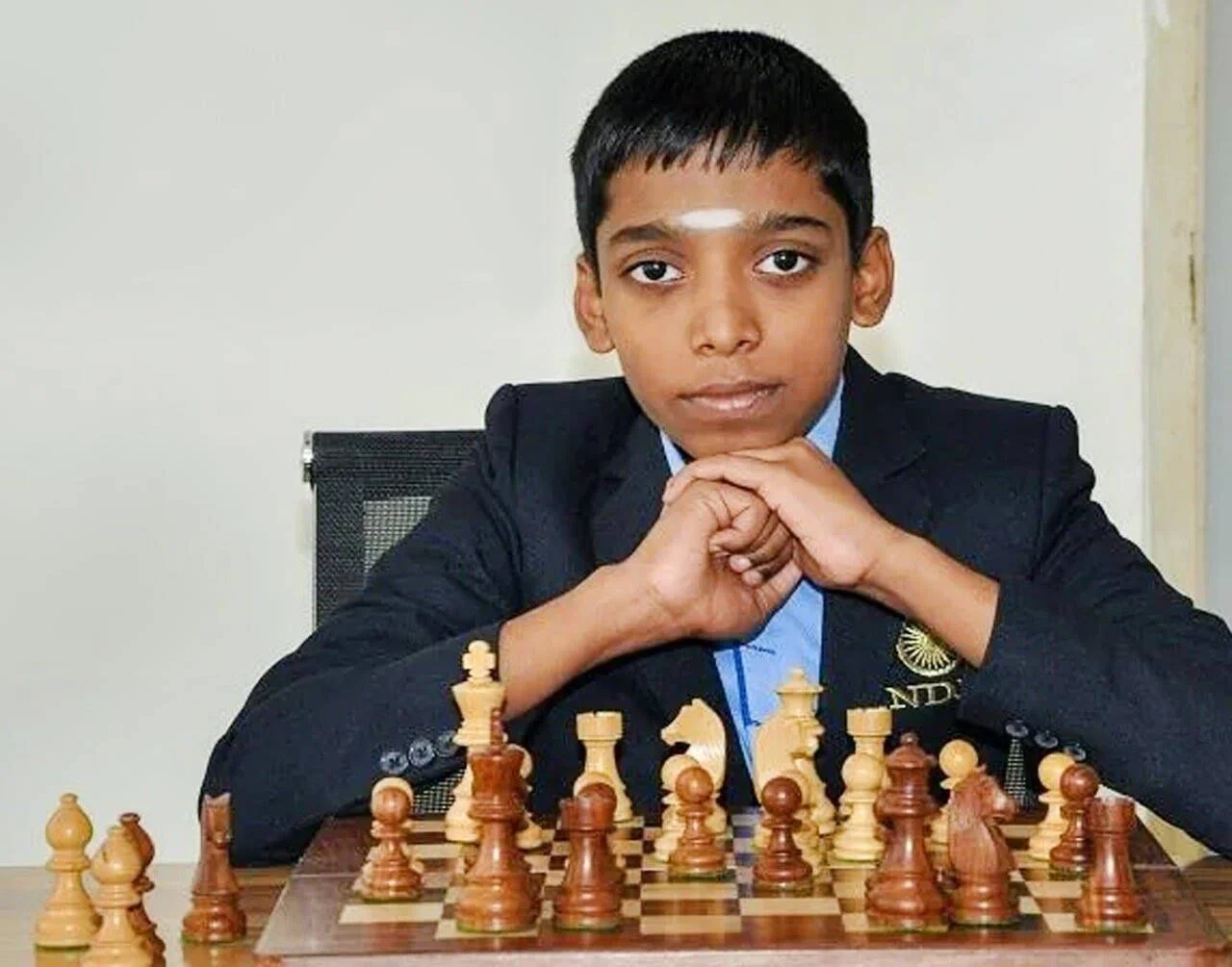 Niño indio con traje y tilak en la frente, apoya las manos bajo el mentón frente a un tablero de ajedrez con piezas dispuestas.