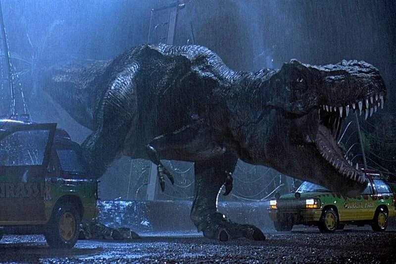 Un Tiranosaurio Rex ruge entre dos vehículos de Jurassic Park durante una tormenta nocturna, en escena icónica de la película.