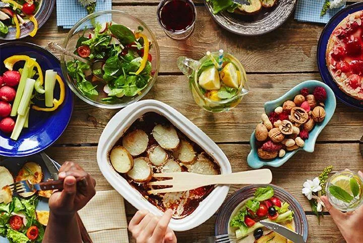 Vista cenital de una mesa de madera con ensaladas, gratén de patatas, frutos secos, bebidas y otros platos mientras dos personas se sirven.
