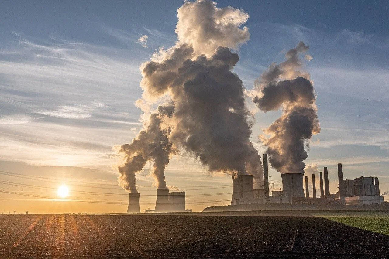 Torres de enfriamiento de una central eléctrica expulsan grandes columnas de vapor y humo al atardecer, sobre campos agrícolas.