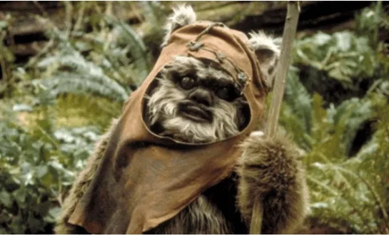 Personaje Ewok con capucha marrón rodeado de vegetación en escena de Star Wars.