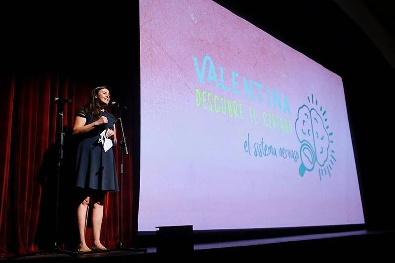 Una mujer habla en un micrófono sobre un escenario frente a una pantalla con el título 'Valentina Descubre el Cerebro - El Sistema Nervioso'.