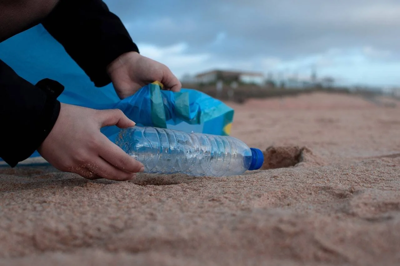 Una persona recoge una botella de plástico de la arena de una playa y la deposita en una bolsa azul de basura.