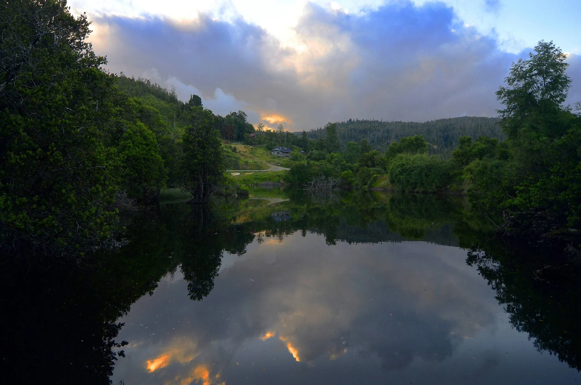 Río tranquilo rodeado de bosque verde refleja nubes iluminadas por el atardecer, con una casa rural al fondo entre colinas.