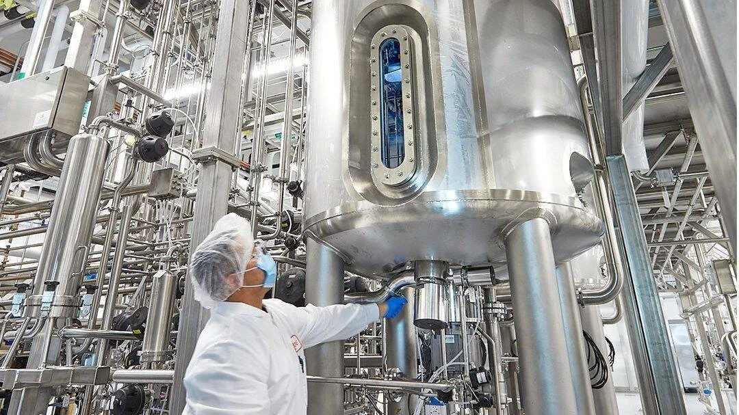 Técnico con bata, mascarilla y cofia opera un gran biorreactor de acero inoxidable en una planta de producción industrial.