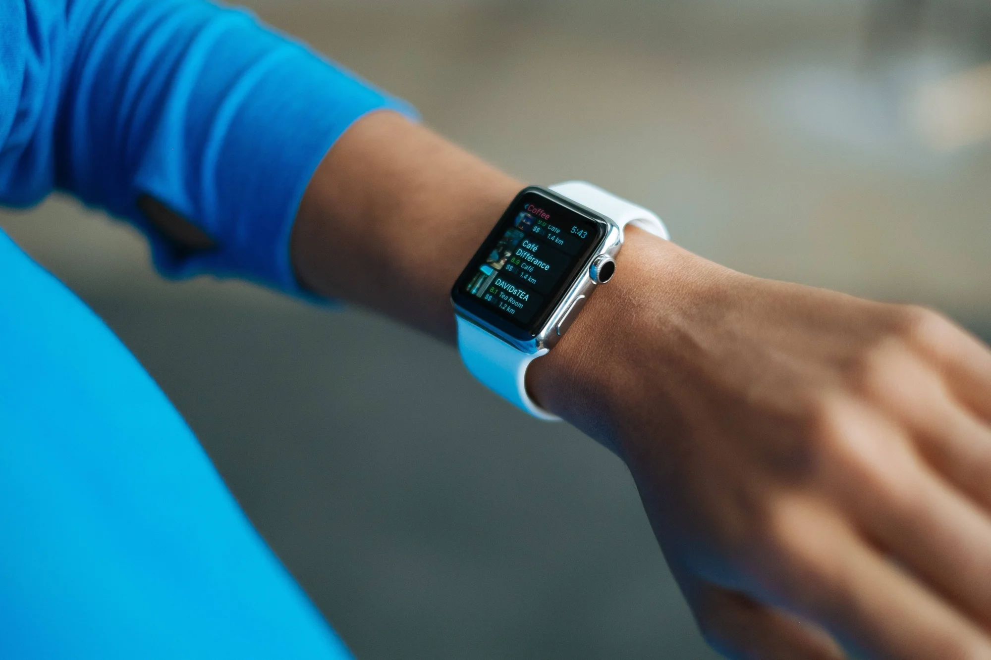 Persona con ropa deportiva azul muestra un smartwatch en su muñeca con una app que lista cafeterías cercanas y sus distancias.