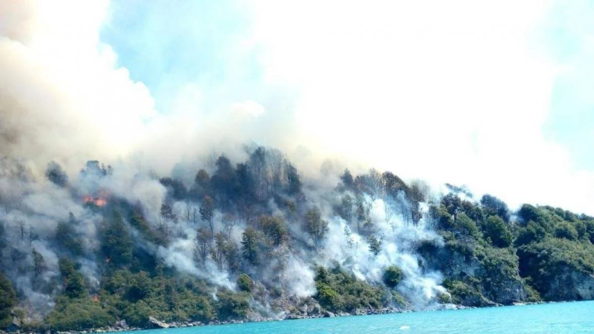 Incendio forestal activo consume árboles en una ladera rocosa a orillas de un lago de aguas turquesas, con llamas y columnas de humo.