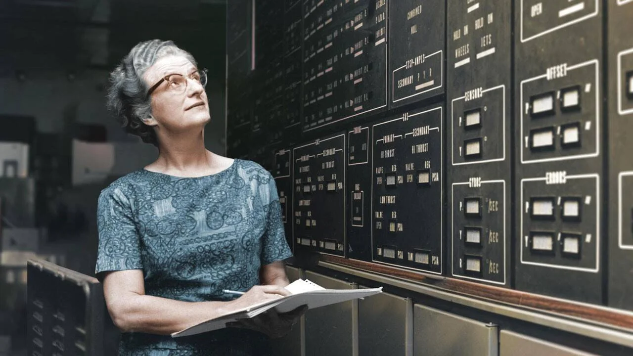 Mujer mayor con gafas sostiene un cuaderno mientras revisa un gran panel de control de instrumentos aeroespaciales.