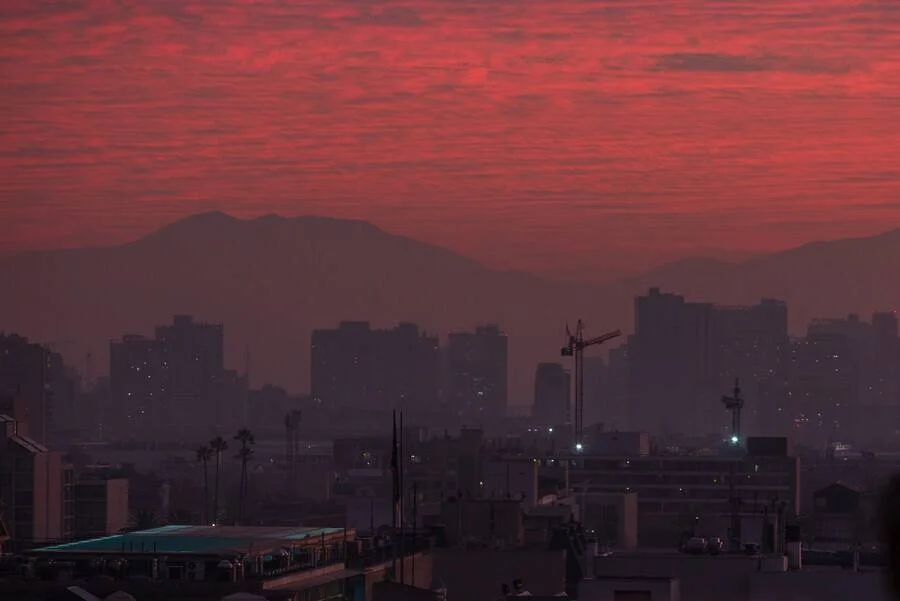 Skyline urbano envuelto en smog bajo un cielo rojo intenso al atardecer, con montañas al fondo.