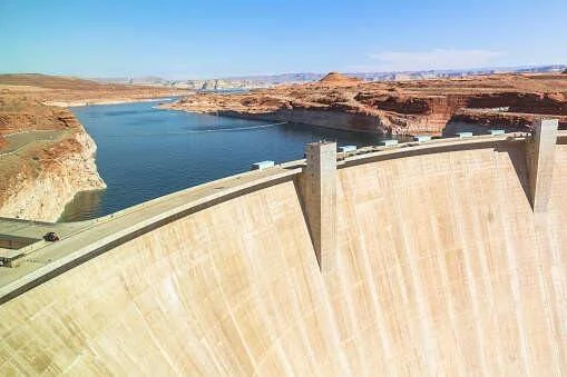 Gran presa de concreto en arco retiene un embalse azul rodeado de acantilados rocosos rojizos bajo cielo despejado.