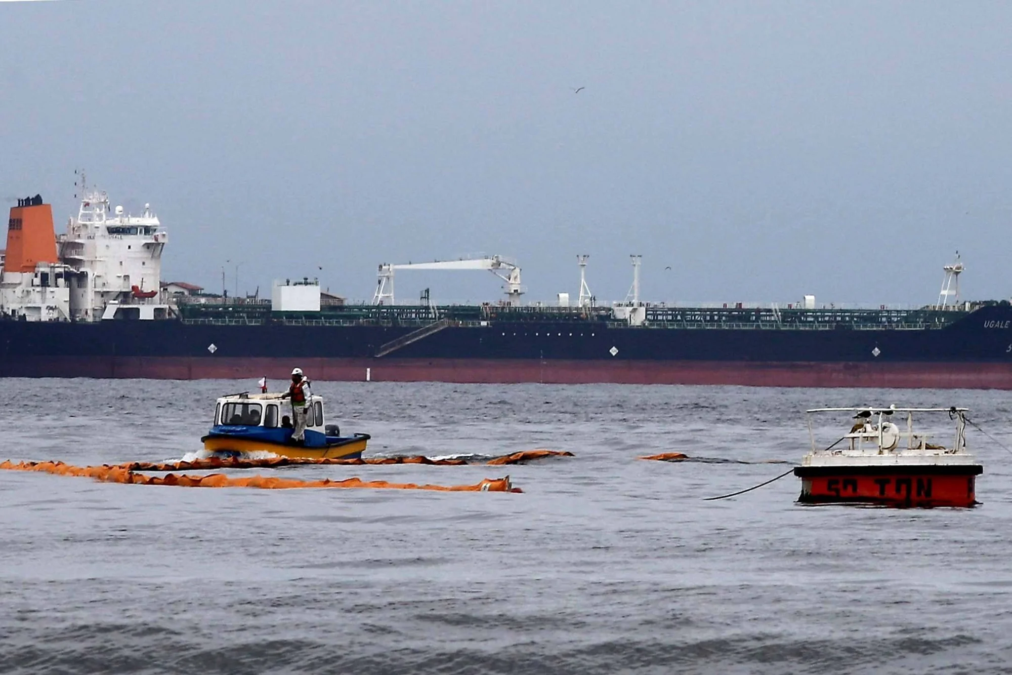 Embarcaciones despliegan barreras de contención naranjas en el mar frente a un gran buque tanque para controlar un derrame de petróleo.