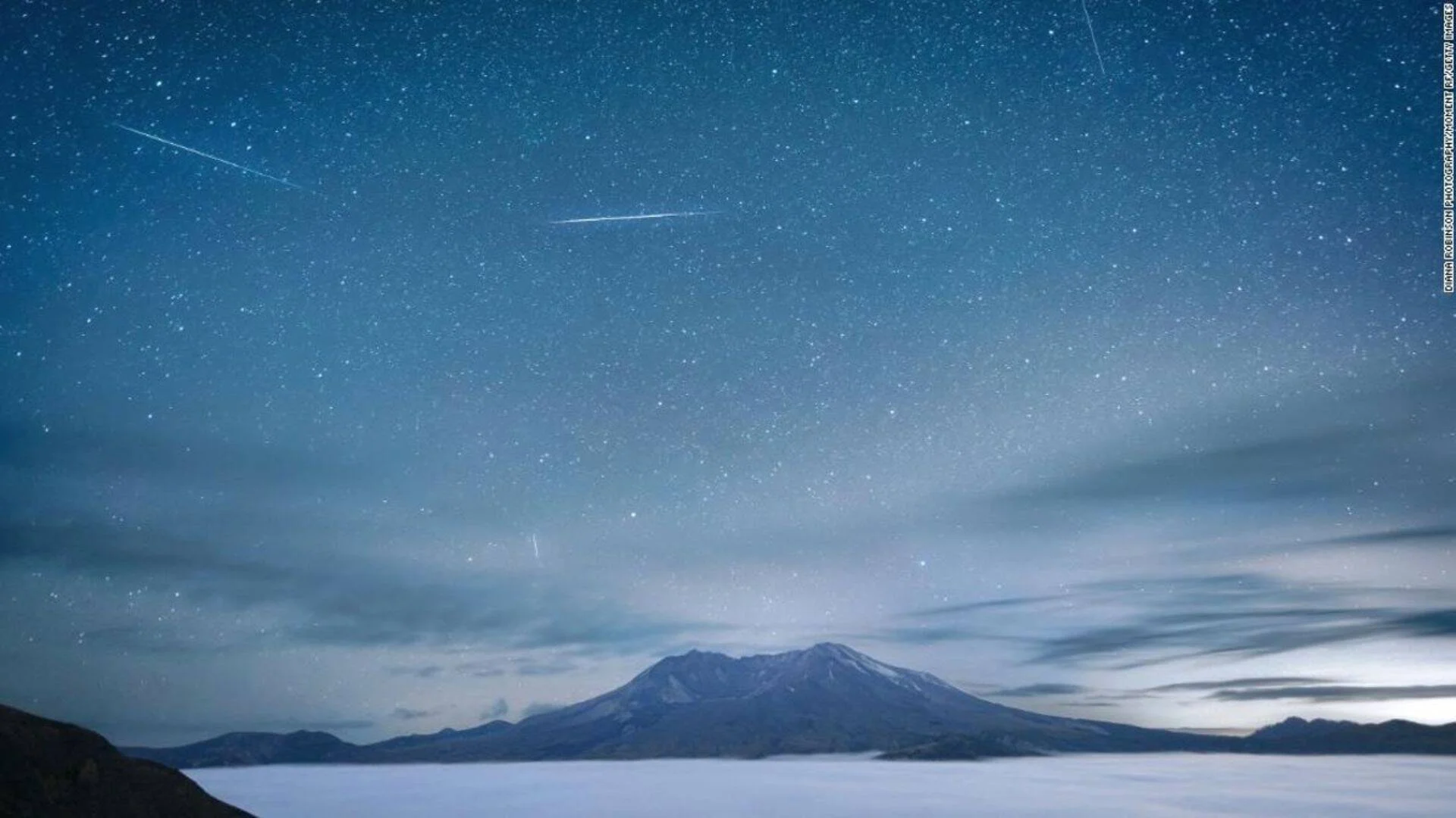 Meteoros cruzando el cielo estrellado sobre un volcán rodeado de nubes bajas durante la noche.