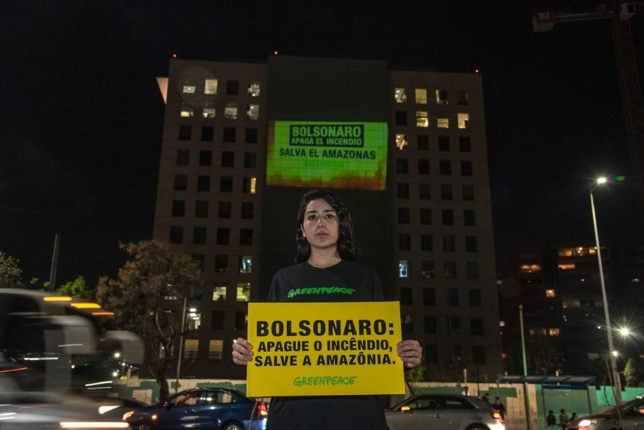Activista de Greenpeace sostiene cartel amarillo exigiendo salvar la Amazonia, con proyección del mismo mensaje en un edificio al fondo.
