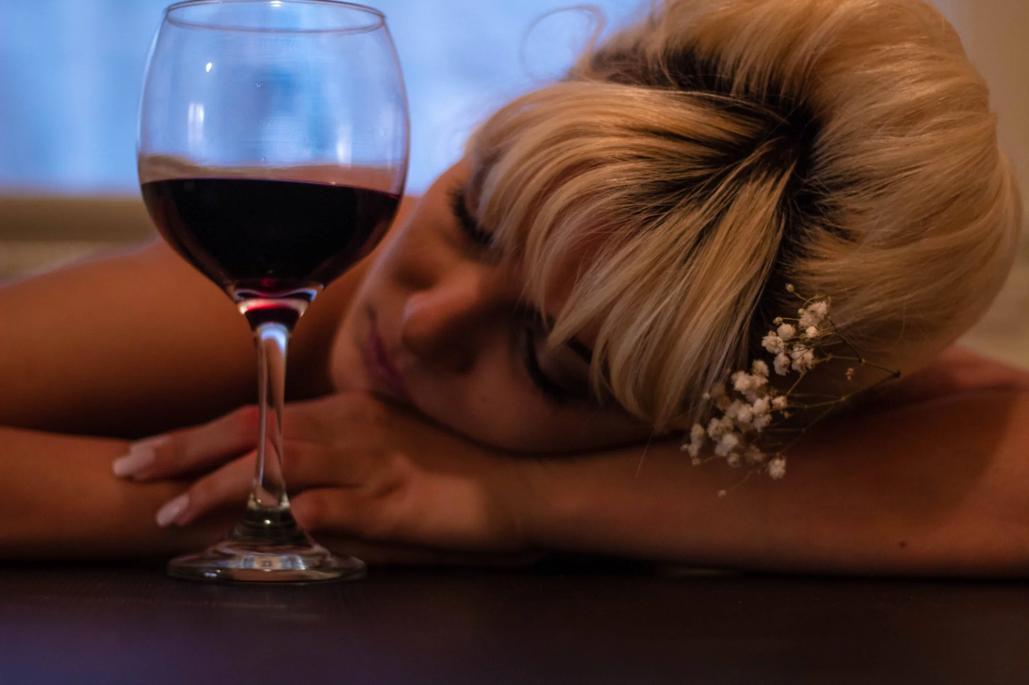 Mujer rubia con flores en el cabello recostada sobre una mesa junto a una copa de vino tinto, aparentando estar dormida o inconsciente.