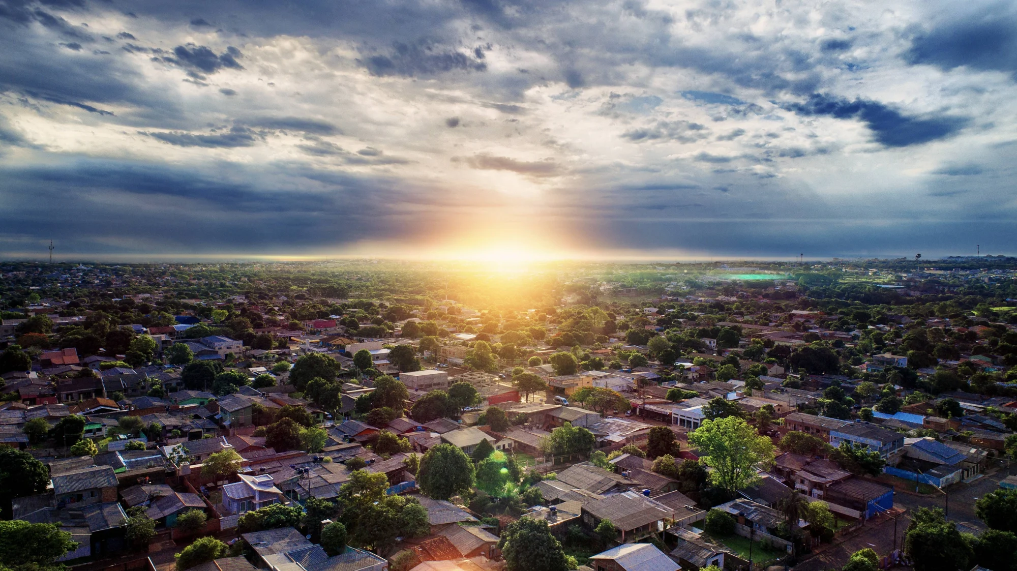 Panorámica aérea de una ciudad de casas bajas y árboles frondosos bajo un cielo nublado con el sol en el horizonte.