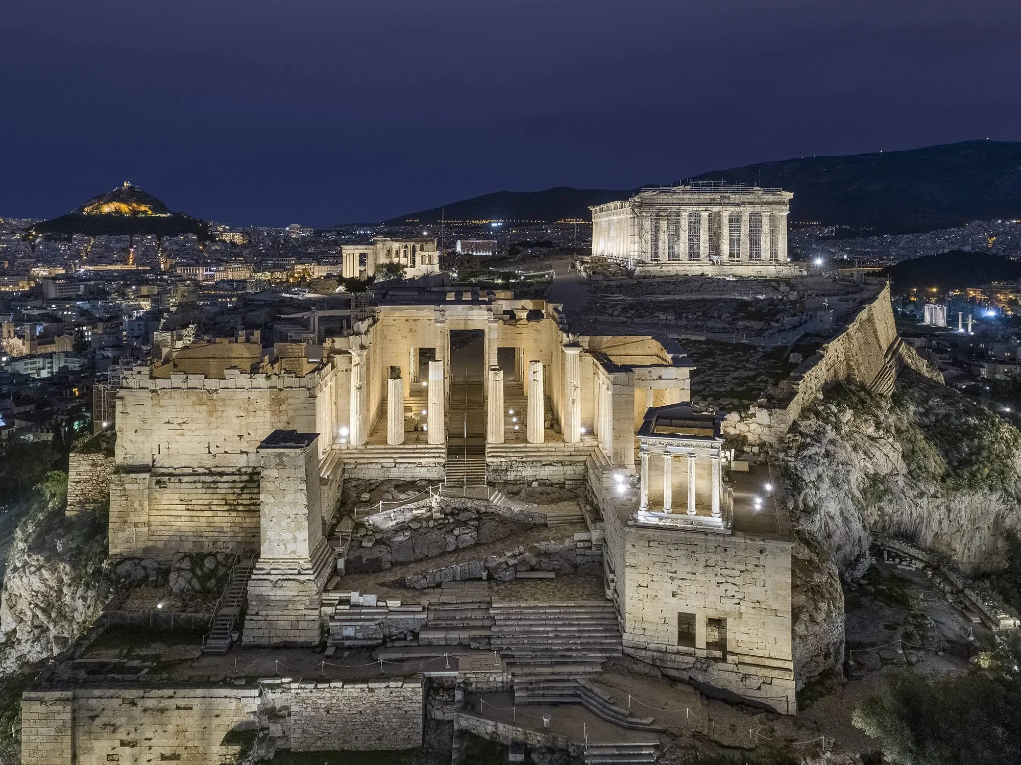 Vista aérea nocturna de los Propíleos y el Partenón iluminados en la Acrópolis de Atenas, con la ciudad extendida al fondo.