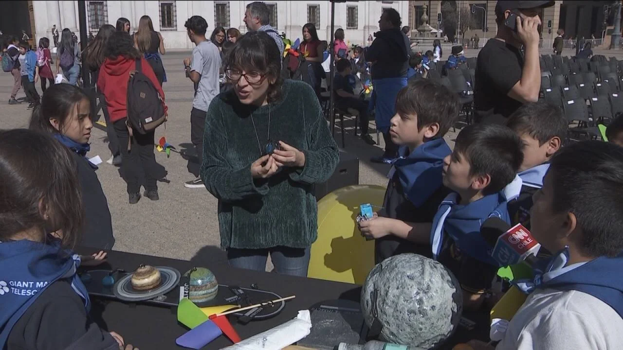 Una mujer explica modelos del sistema solar a niños escolares en una actividad educativa al aire libre en una plaza.