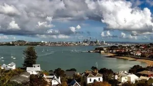 Vista panorámica de una bahía con veleros, barrio residencial en primer plano y skyline urbano al fondo bajo cielo nublado.
