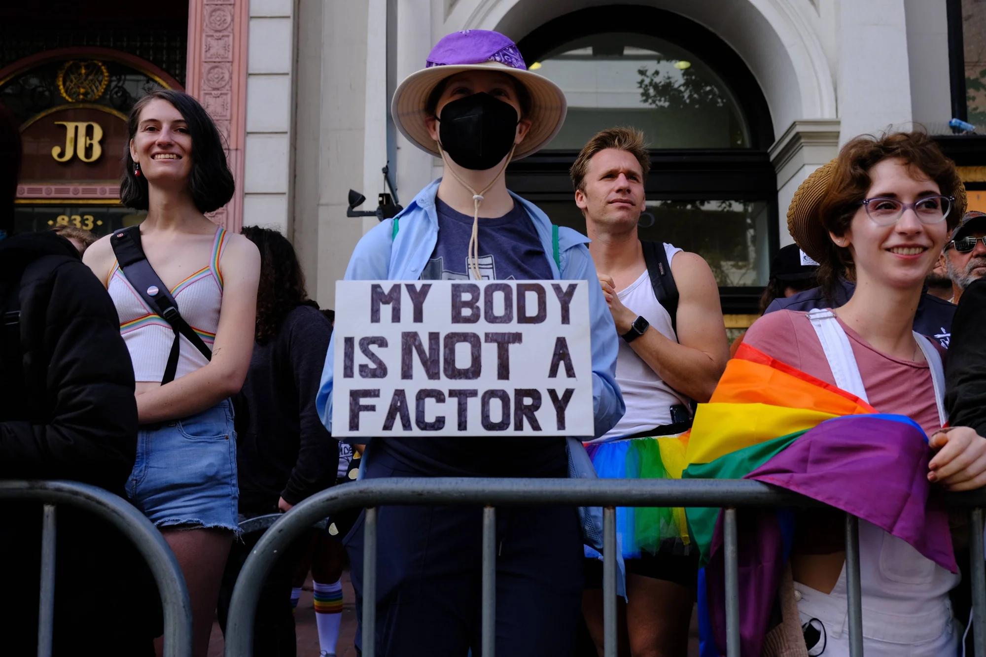 Manifestantes en una protesta; uno sostiene un cartel con "My body is not a factory" y otro lleva una bandera arcoíris LGBTQ+.
