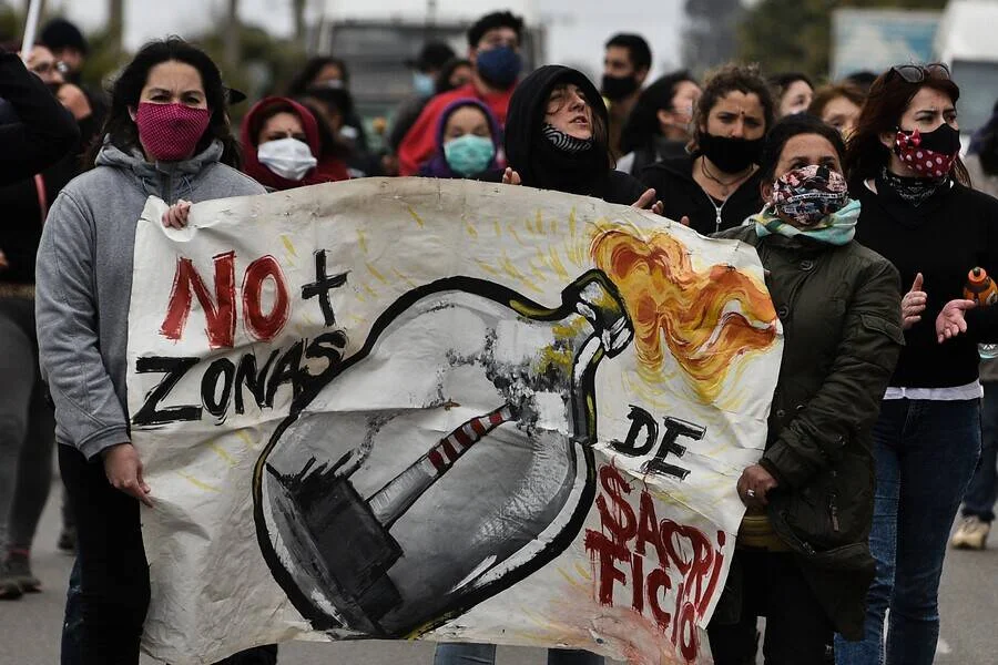Personas con mascarillas sostienen un cartel pintado que dice "No más zonas de sacrificio" durante una protesta ambiental.