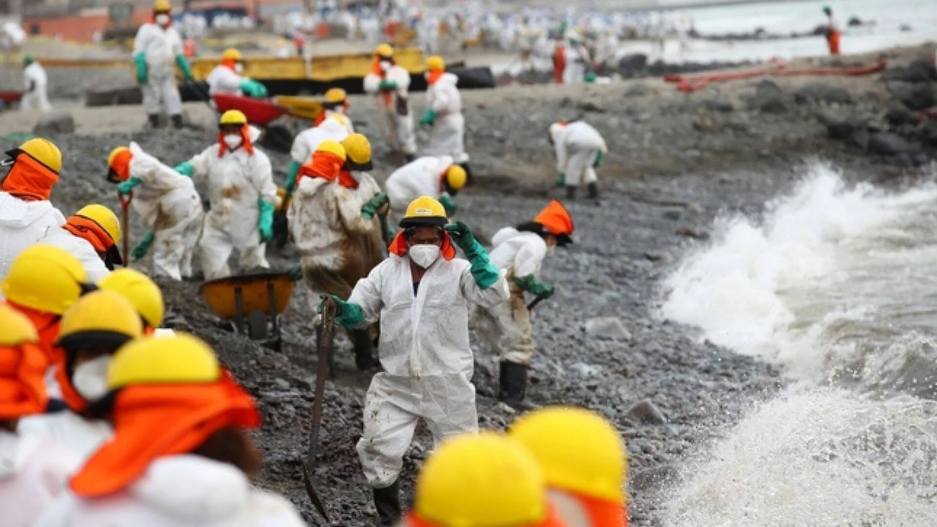 Decenas de trabajadores con trajes blancos de protección y cascos amarillos limpian manualmente una costa rocosa contaminada por petróleo.