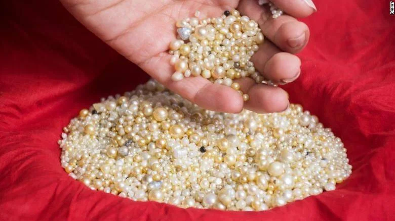 Una mano sostiene un puñado de perlas de distintos tamaños y colores sobre una gran colección de perlas apoyada en tela roja.