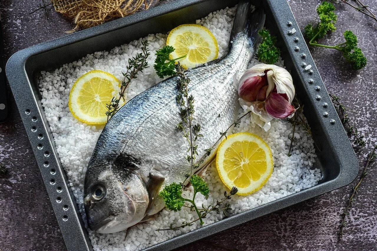 Dorada entera cruda sobre una cama de sal gruesa en bandeja de horno, acompañada de rodajas de limón, ajo y hierbas frescas.