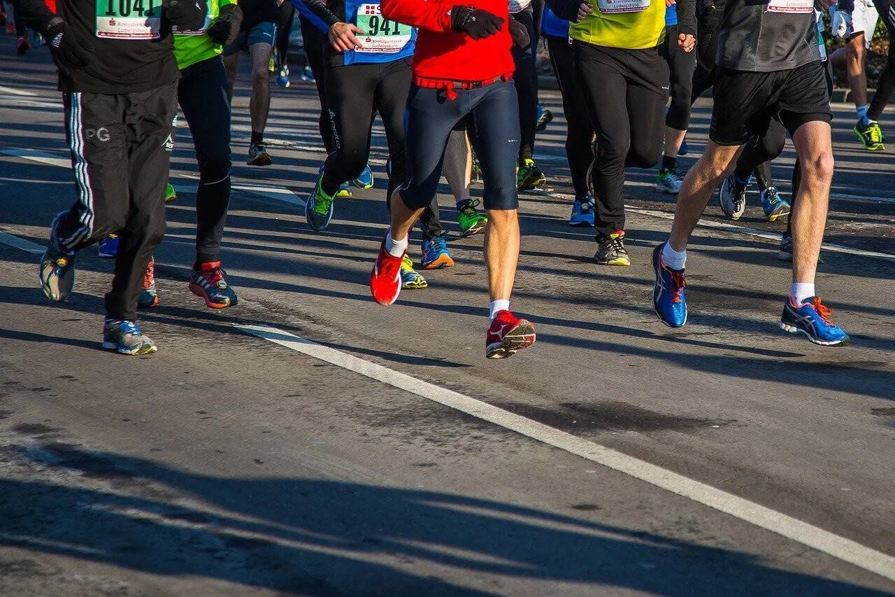 Grupo de corredores con dorsales compitiendo en una carrera popular sobre asfalto, vistos desde la cintura hacia abajo.