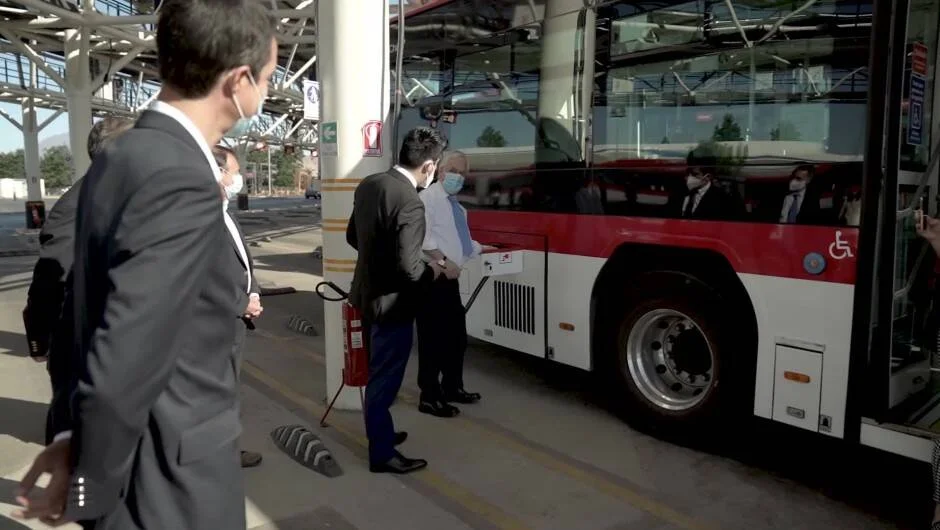 Hombres de traje y mascarilla inspeccionan un autobús rojo y blanco en una estación de transporte público.