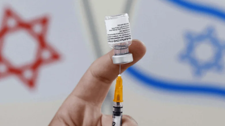 Mano sostiene una jeringa con vial de vacuna COVID-19 ante una bandera de Israel desenfocada al fondo.