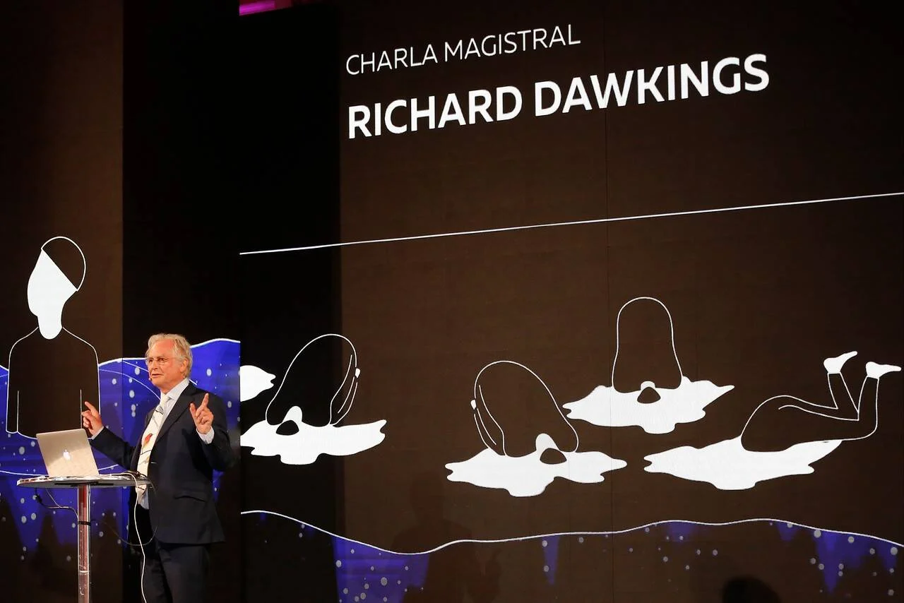 Richard Dawkins gesticula frente a una pantalla que anuncia su charla magistral, con ilustraciones de evolución humana al fondo.