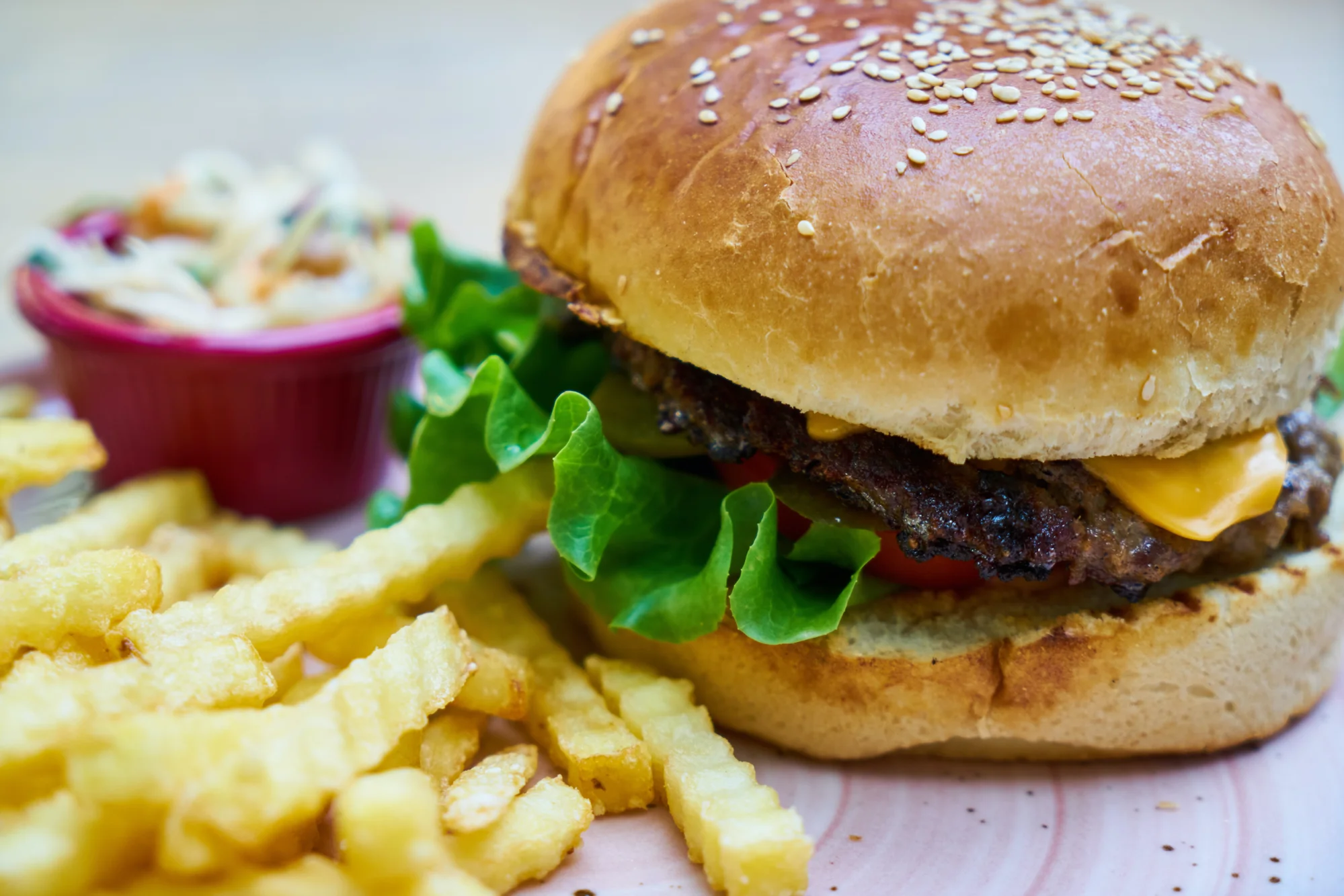 Hamburguesa con cheddar, lechuga y tomate en pan de sésamo, acompañada de papas fritas y un recipiente de coleslaw.