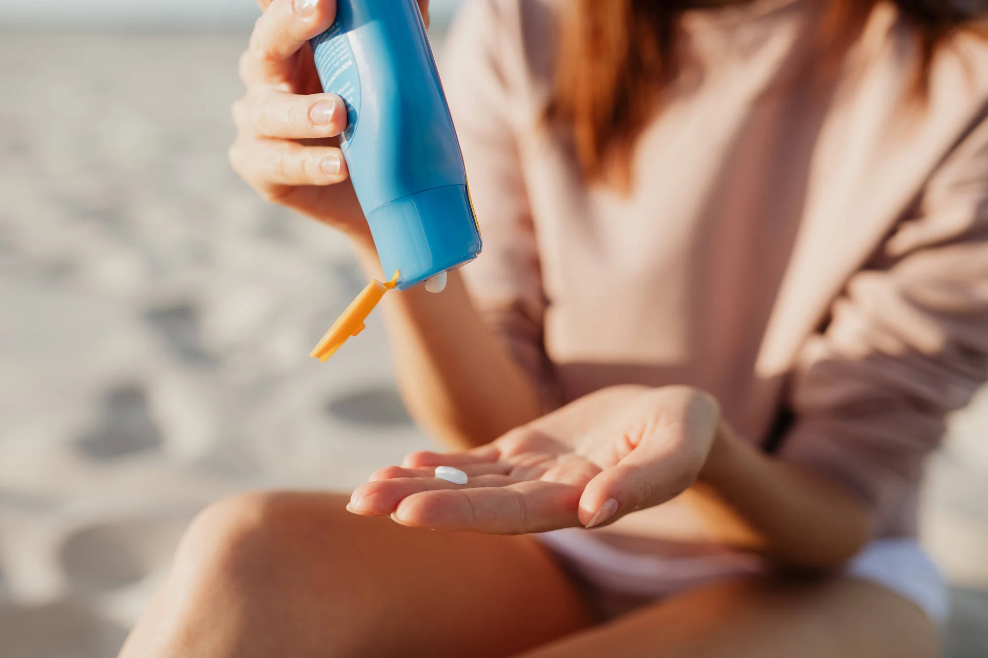 Una mujer dispensa crema protectora solar de un envase azul sobre su mano, sentada en la playa.