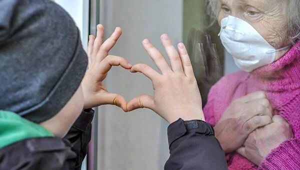 Un niño forma un corazón con sus manos en el vidrio ante una anciana con mascarilla, separados por una ventana.