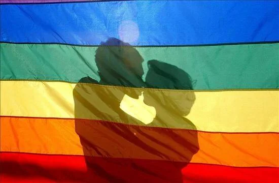 Silueta de dos personas besándose proyectada sobre una bandera del orgullo LGBT con franjas de colores.
