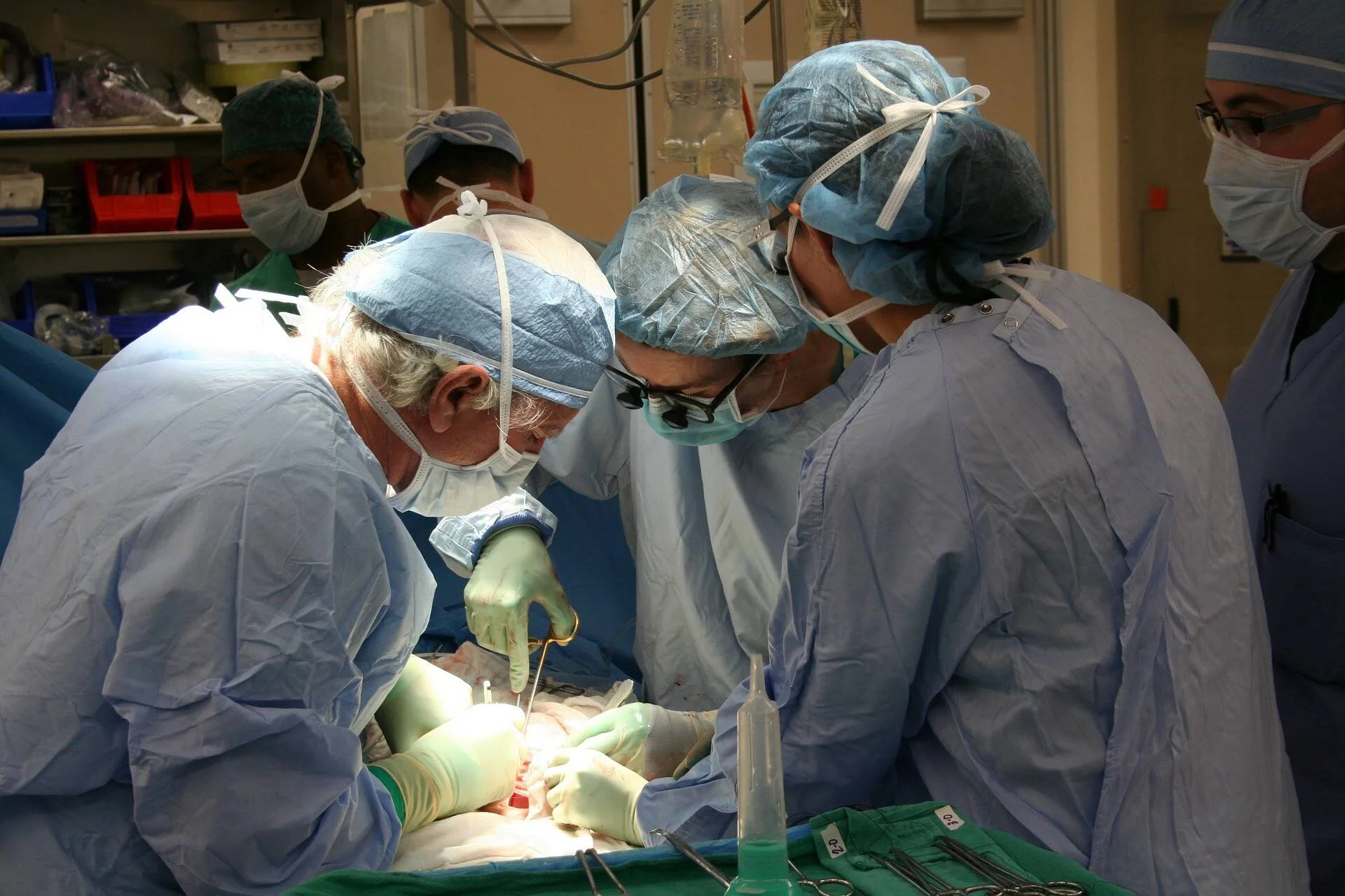Varios cirujanos con bata, gorro y mascarilla quirúrgica operan a un paciente en un quirófano iluminado.